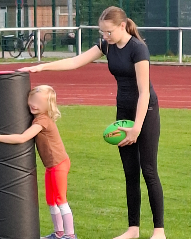 Kea hält einen Rugbyball und stabilisiert eine Tacklesäule, die ein Kind beim Training der Rugby Flöhe umfasst.