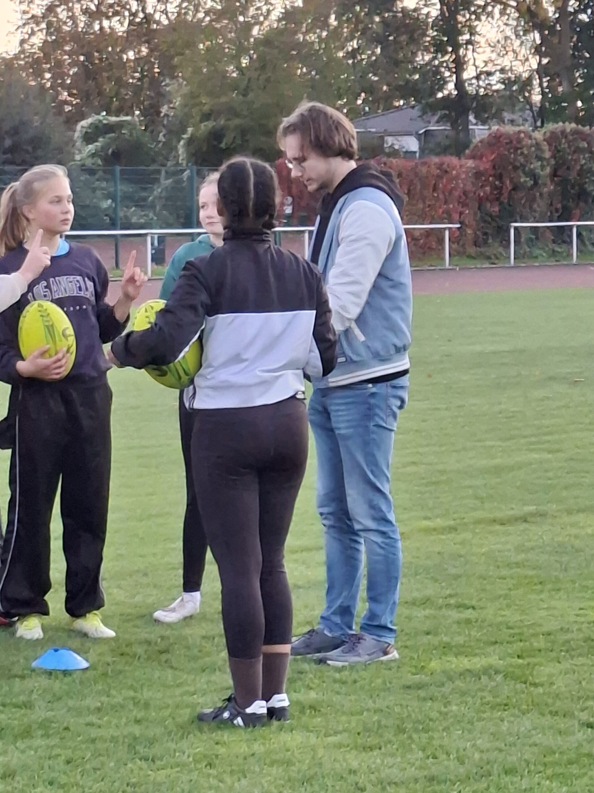 Trainer Jannis steht mit drei Spielerinnen der Rugby Foxes auf dem Trainingsplatz, zwei halten Rugbybälle.